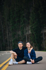 Positive man and woman tourists on vacation sat down to rest on the asphalt road in the mountains, hugging and smiling against the backdrop of a coniferous forest