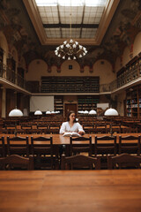 Photo of a woman in a white blouse sitting at a table in a beautiful public library and reading a book.