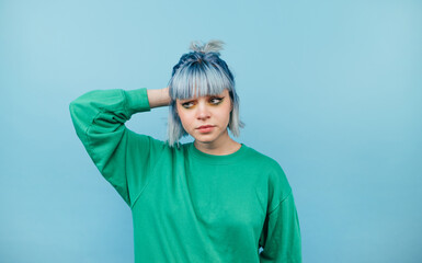 Beautiful woman in colored casual clothes and blue hair stands on a blue background and looks suspiciously to the side on copy space