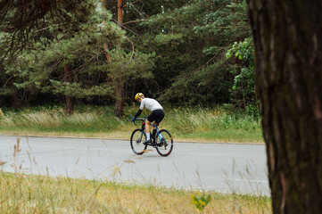 A man in a bicycle ride rides on an asphalt road in the woods and looks at the camera.