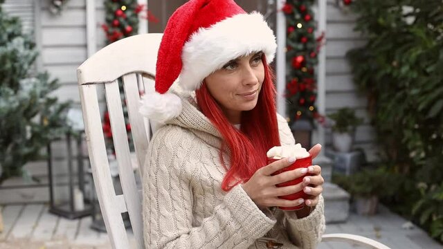 Beautiful hipster girl with red hair in a santa claus hat sits in a rocking chair on the veranda of the house, enjoys the winter holidays and christmas mood and drinks cocoa with marshmallows 
