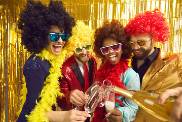 Group of joyful diverse friends in funny Afro wigs drinking champagne from flutes at groovy retro style disco party. Happy caucasian and black African American people having fun and enjoying nightlife