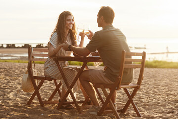 Full length portrait of young couple drinking champagne on beach while enjoying romantic date in sunlight, copy space