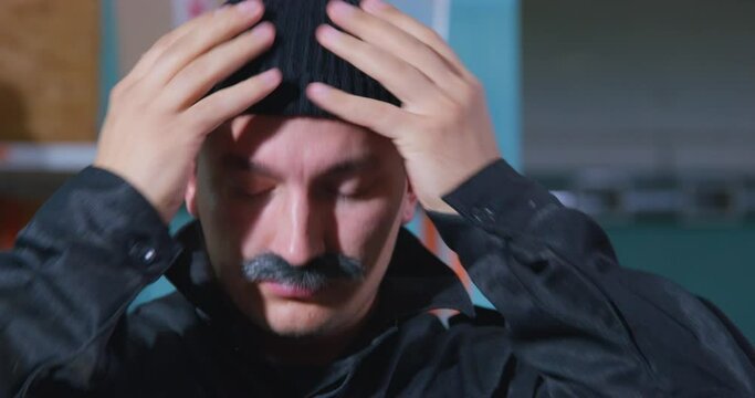 Suspicious looking man with gray mustache puts on warm woolen black hat, because he is going to go outside, and it is cold there, front view. Sudden cold snap before the heating season.