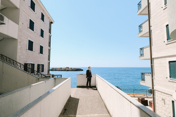 Elegant man in a black suit stands on a terrace between two buildings above the sea and looks into the distance