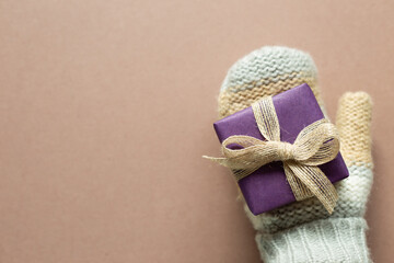 Hand in winter knitted gloves with purple gift box on brown background. anniversary, christmas, greeting concept. top view, copy space