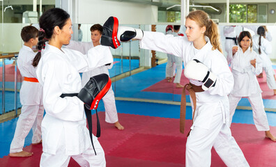 Obraz premium Oriental female trainer in kimono and focus mitts teaching young girl jabbing during group karate class. Other kids exercising in background.