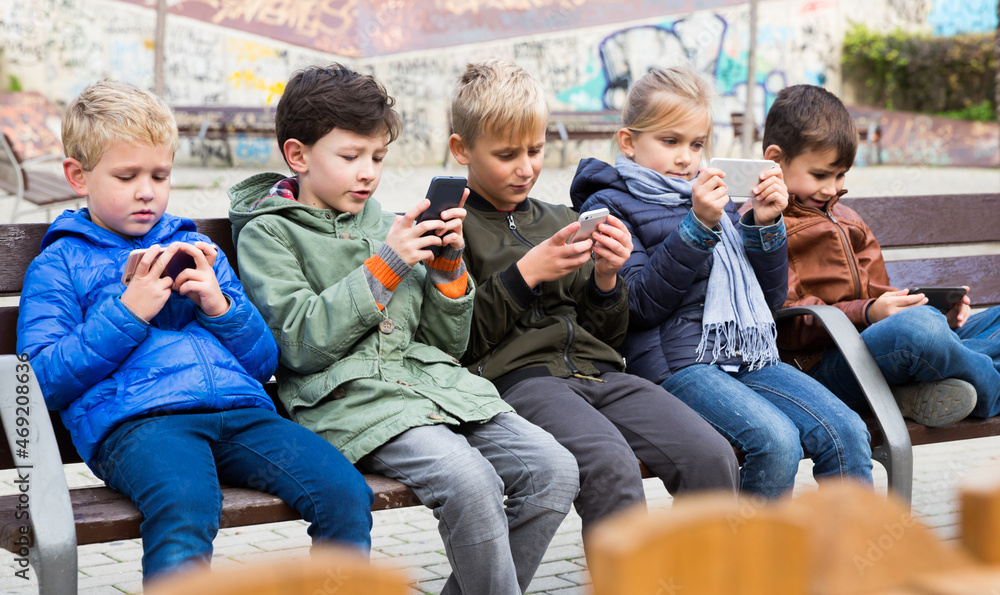 Wall mural group of preteen children sitting on bench outdoors, addicted in their phones..