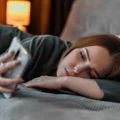 Sad teenage girl suffering from insomnia surfing Internet or chatting using smartphone at night lying on bed. Depression Internet addiction. Young woman depression closeup square portrait