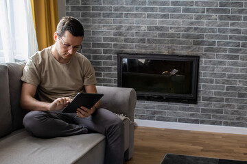 A young man is using a digital tablet. A guy is typing while sitting on a sofa