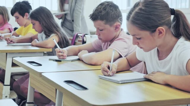 Upset serious preteen boy and girl listening to teacher and writing exercises in notebook at lesson in primary school