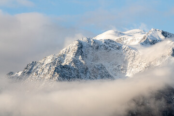 Snow capped mountain peaks in northern Canada with misty, foggy clouds covering the peaks with white, winter theme and spectacular blue sky behind. 