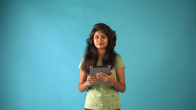 A young Indian girl in orange frock standing with a tab looking at the camera in an isolated blue background studio.
