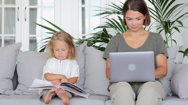 Mother and daughter work and study at home. spbi Pretty short haired woman freelancer uses modern laptop while little girl looks at pictures in book in living room