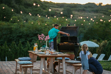 Young adult man barbecuing while his wife and daughter is playing with dog