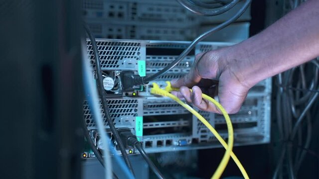 Engineer IT technician sets up system. African-American man switches colorful cables spbas to run checkup of server equipment on rack in data center closeup