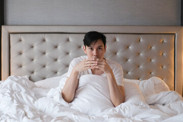 Young male sitting on bed with cup of coffee in the morning after getting up. Handsome young man resting sitting in his bed. Young man in white t shirt feeling comfortable and sitting on bed.