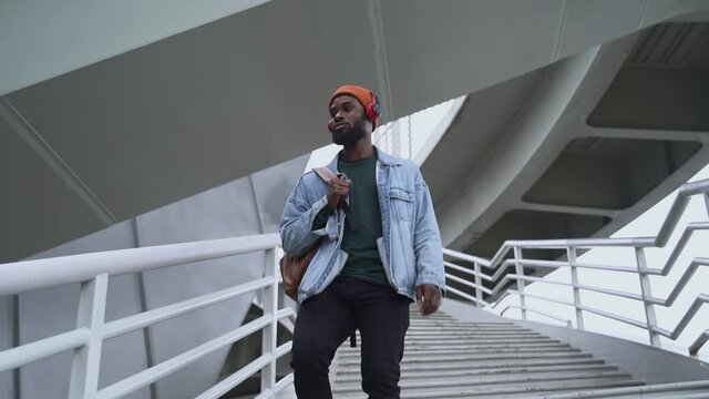 Contemporary gadgets to enjoy audio. Tranquil African-American guy in casual clothes with earphones spbas and backpack goes down pedestrian bridge stairs outdoors