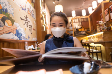 Asian woman wear medical mask and looking at menu in sushi restaurant. Food and social issues concept