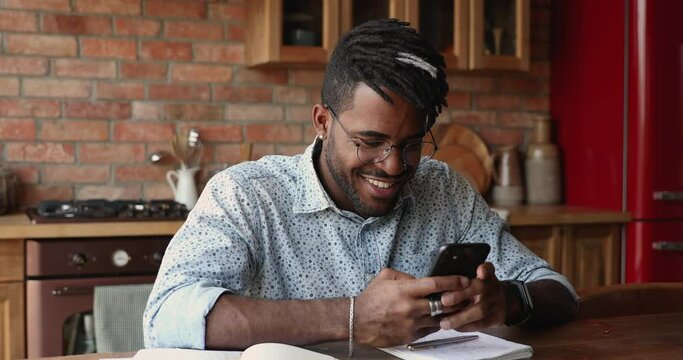 Handsome African hipster guy in glasses and dreadlocks hairstyle sit in kitchen holds smartphone chatting, watch received video, listen favourite band music on-line. Fun, leisure, modern tech concept