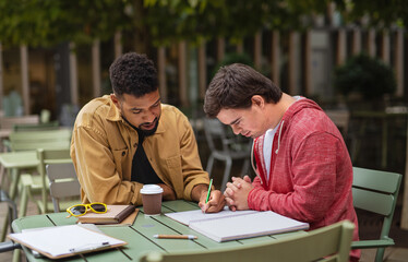 Young man with Down syndrome with his mentoring friend sitting outdoors in cafe and studying.