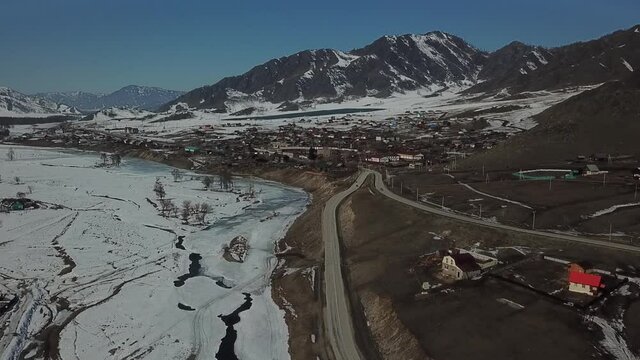 Aerial shot in movement of mountains with melting snow on it and melting river ice near village when spring is coming. Some houses and asphalt roads also