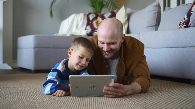 Father and toddler son have fun. Careful happy bald and bearded parent and spbd adorable little boy enjoy videogame on tablet computer lying on floor at home