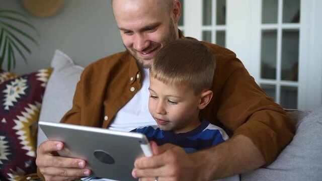 Father spends time with little son. Happy smiling bald man with adorable toddler boy watch movie via tablet sitting on couch in living room closeup spbd