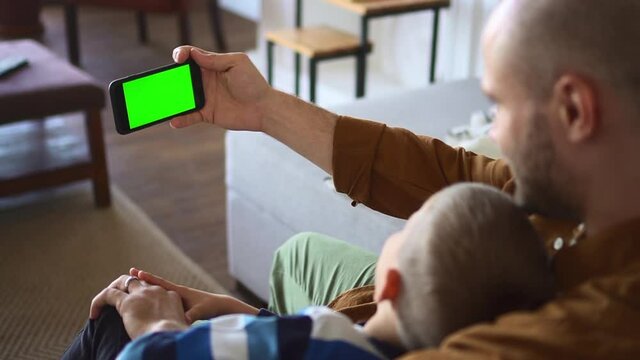 Father and son at videochat. Happy little child and young parent talk to grandma via cellphone with chroma key display sitting on sofa closeup spbd