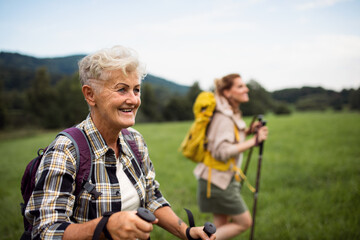 Happy mid adult woman with trekking poles hiking with active senior mother outdoors in nature.