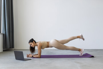 Fototapeta na wymiar Young woman exercising at home in a living room. Video lesson. Young woman repeating exercises while watching online workout session. Beautiful young woman doing fitness exercise at home