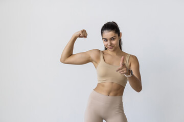Obraz premium Sporty woman demonstrating biceps and points her finger at you, isolated on white background.
