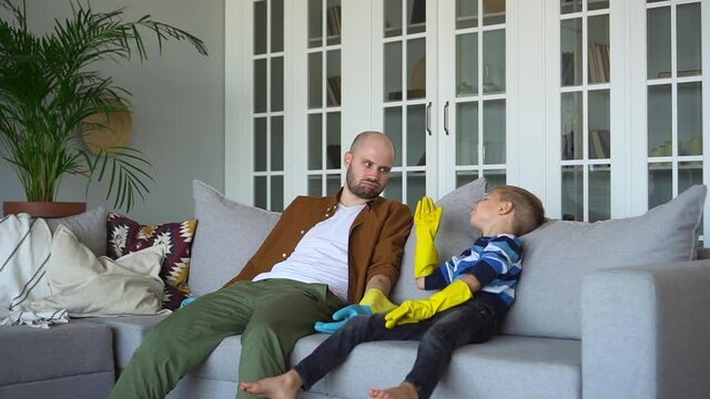 Father and toddler do housework. Tired preschooler son and parent wearing rubber gloves fall down on sofa and give high five after finished cleaning at home spbd