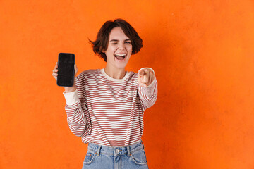 Young woman showing cellphone and pointing finger at camera