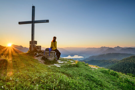 Female hiker sitting by summit cross on mountain peak at sunrise, Carinthia, Austria