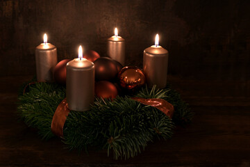 Small Advent wreath with four lit copper colored candles and Christmas decoration baubles against a...