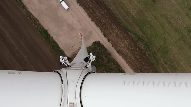 Service engineers with safety harnesses and long ropes go down wind turbine propeller tower above agricultural plowed field upper view