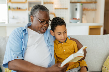 Grandfather hugging his beloved grandson and reading a story to him while sitting on the sofa at home. - 469253446