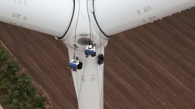 Skilled technician fitters with safety harnesses go down wind turbine pylon blade using long ropes above agricultural plowed field aerial view
