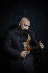 Stylish handsome man with a black beard in a black suit with a naked torso and hairy chest stands with a beautiful rose in his pants