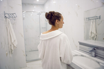 woman standing in the bathroom next to the shower cabin in hotel apartment in a white robe
