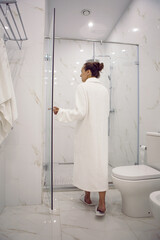 woman standing in the bathroom next to the shower cabin in hotel apartment in a white robe