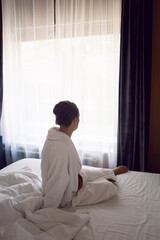 woman in a white robe is sitting on a bed by the window in hotel apartment after a shower