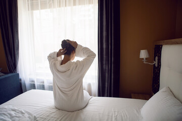 woman in a white robe is sitting on a bed by the window in hotel apartment after a shower