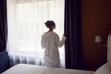 Fototapeta na wymiar woman in a white robe is stand on a bed by the window in hotel apartment after a shower
