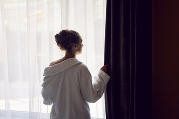 woman in a white robe is stand on a bed by the window in hotel apartment after a shower