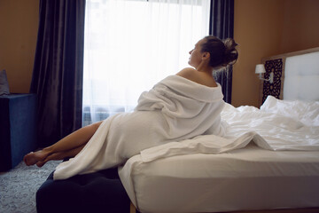 woman in a white bathrobe is sitting on the edge of the bed and drinking tea from mug at a large window in the hotel