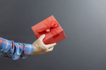 The hand of a man in a plaid shirt holds a red box with a festive gift on a gray background