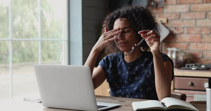 Exhausted African freelancer woman sit at table takes off glasses, resting from laptop work, having dry irritable eyes after long modern tech usage. Conjunctivitis, asthenopia, ocular fatigue concept