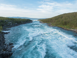 Fototapeta premium Blue wild water of Vuojatadno river rapids and mouth to Akkajaure lake, rocks and birch tree forest in the background. Summer Lapland Sweden arctic landscape.